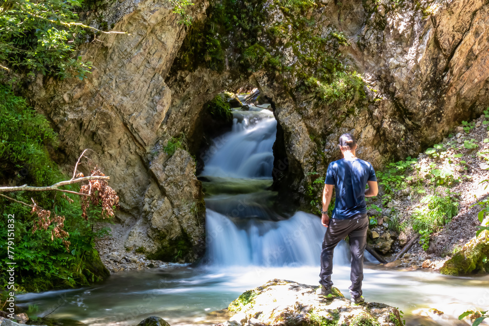 Man standing on rock with scenic view of majestic waterfall cascade of Bodentaler Felsentor in Tscheppaschlucht, Loibl Valley, Karawanks, Carinthia, Austria. Water cascading down cliff face into pool Man standing on rock with scenic view of majestic waterfall cascade of Bodentaler Felsentor in Tscheppaschlucht, Loibl Valley, Karawanks, Carinthia, Austria. Water cascading down cliff face into pool