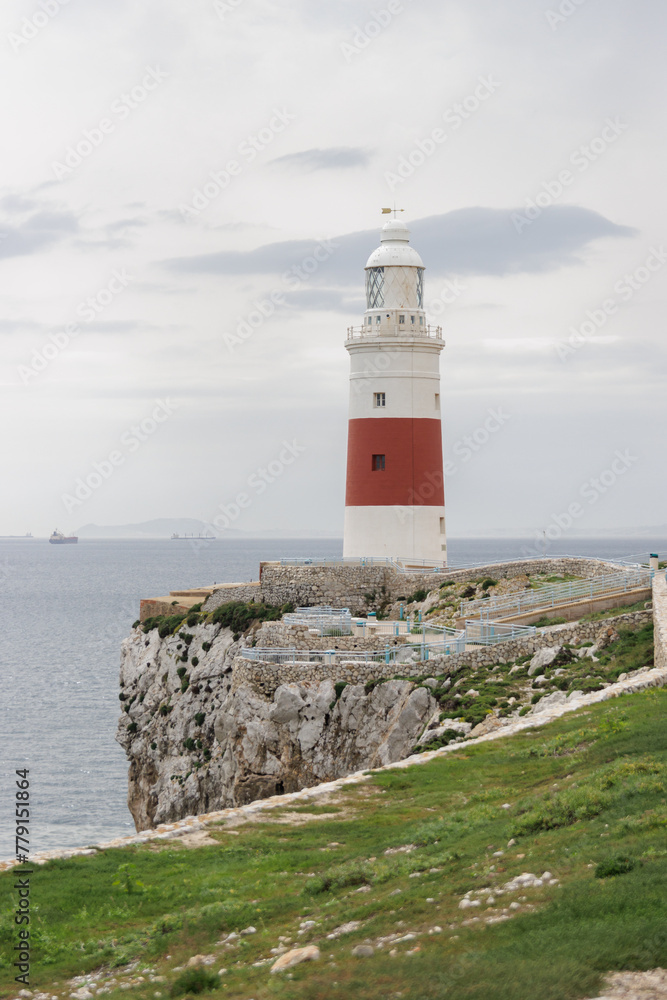 A lighthouse is on a rocky cliff overlooking the ocean. The lighthouse is red and white. The ocean is calm and the sky is cloudy