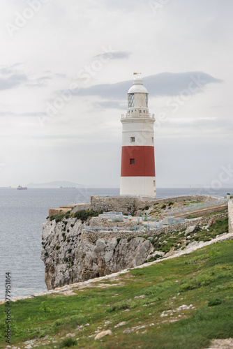 A lighthouse is on a rocky cliff overlooking the ocean. The lighthouse is red and white. The ocean is calm and the sky is cloudy