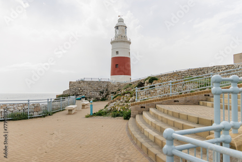 A lighthouse is on a rocky shore with a blue railing. The lighthouse is red and white