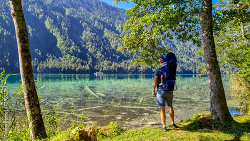 Man with baby carrier looking at scenic view of east bank of alpine lake Weissensee, Gailtal Alps, Carinthia, Austria. Bathing lake surrounded by mountains of Austrian Alps. Untouched nature in summer