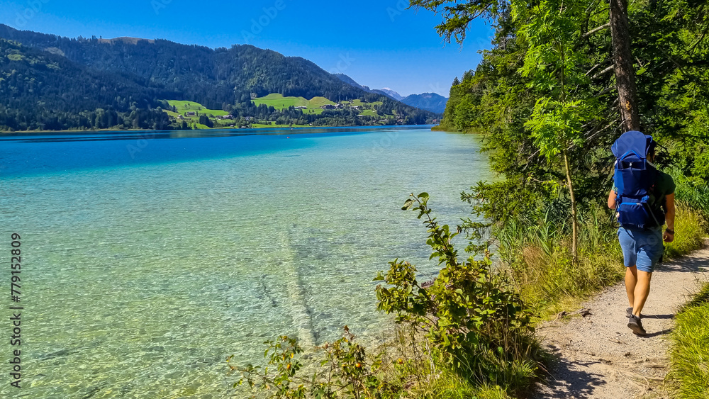 Man with baby carrier looking at scenic view of east bank of alpine lake Weissensee, Gailtal Alps, Carinthia, Austria. Bathing lake surrounded by mountains of Austrian Alps. Untouched nature in summer