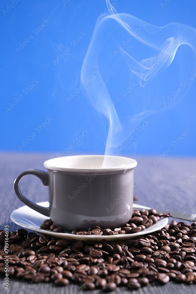 Obraz premium Tazza grigia con piattino bianco, chicchi di caffè sul tavolo e sul piattino, tazza fumante con caffè bollente, tavolo in pietra e sfondo azzurro