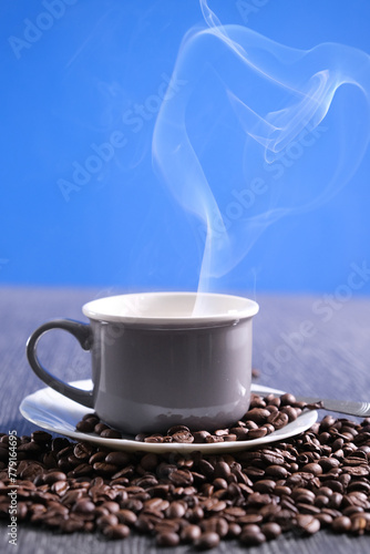 Tazza grigia con piattino bianco, chicchi di caffè sul tavolo e sul piattino, tazza fumante con caffè bollente,  tavolo in pietra e sfondo azzurro