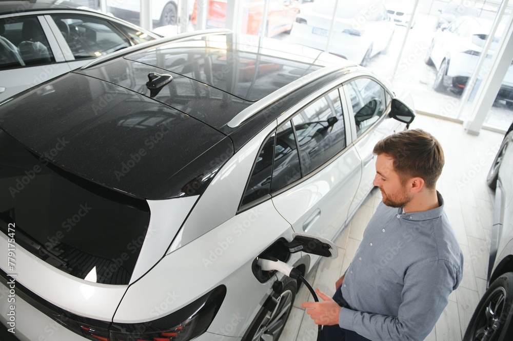 Beard man trying a new charging cable with a car charging station at the motor dealership. Concept of buying electric vehicle. Smart ecological living Beard man trying a new charging cable with a car charging station at the motor dealership. Concept of buying electric vehicle. Smart ecological living