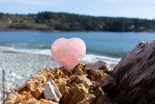 A peaceful image of a beautiful rose quartz crystal heart on a driftwood log with a rocky beach and blue ocean waters in the background.