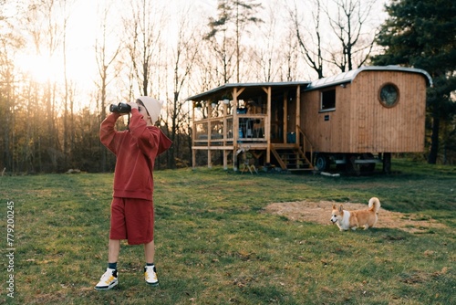 A teenage boy in bright clothes and a hat looks at us through binoculars, a corgi dog next to him photo