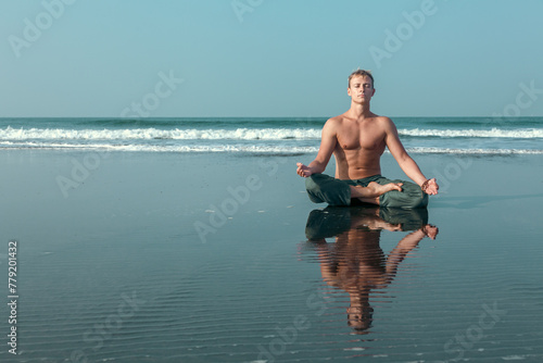 A shirtless young man sits in a meditative lotus pose on the wet sand of a beach, with the ocean waves crashing behind him.