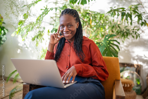 Joyful african american woman smiling looking at camera with laptop on knees siting on armchair surround by indoor plants. Pleased black female freelance working in cozy coworking space with computer.