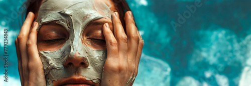 A woman's face covered with a grey mud mask, fingertips touching temples, symbolizes relaxation and skincare. Banner. Copy space photo
