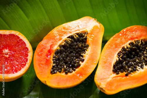 Exotic fruits background. Papaya and orange citrus fresh fruits on tropical leaf green background with shadows of palm tree. Halved fresh organic Papayas and grapefruit exotic fruits close up Top view