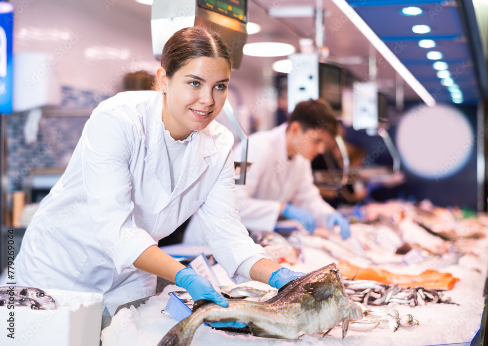 Smiling salesgirl in work clothes holding in hands cod fish in fish shop