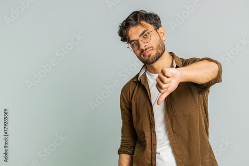 Dislike. Upset unhappy Indian man showing thumbs down sign gesture, expressing discontent, disapproval, dissatisfied negative feedback. Attractive bearded guy indoors isolated on gray background