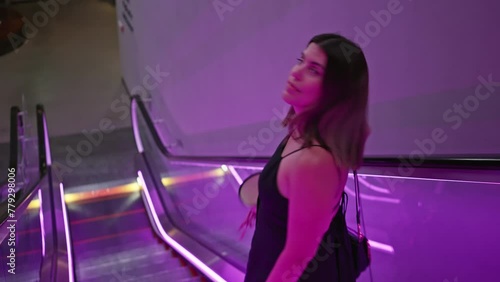 A smiling young woman in a black dress standing on a vibrant lit escalator indoors. photo
