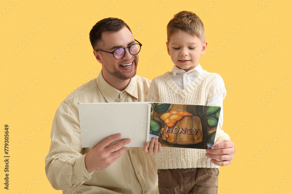Happy father and his little son reading book together on yellow background - obrazy, fototapety, plakaty 