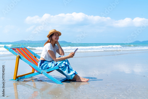 Happy Asian woman enjoy outdoor lifestyle travel ocean on summer holiday vacation. Attractive girl relaxing and using smartphone for online shopping or chatting at tropical island beach in sunny day.