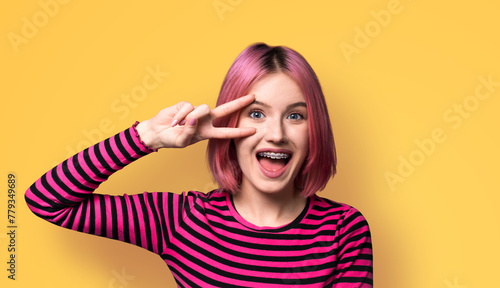 Photo image - happy pink beautiful woman in braces brackets showing v-sign victory peace hand gesture two fingers, opened mouth, isolate yellow wall background. Girl at studio. Dental care ad concept © vgstudio