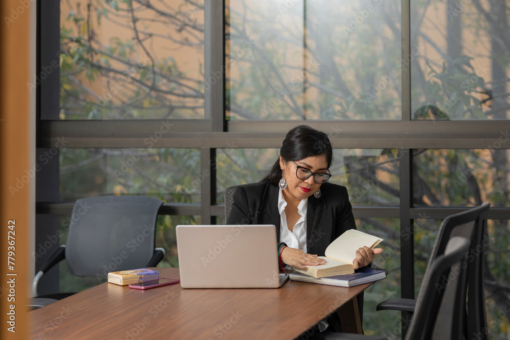 a business woman reading a book in the office during a work day, sitting at her desk with her laptop