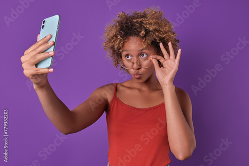 Young beautiful flirty African American woman takes selfie on phone and takes glasses off eyes posing for good photo for social networks or mobile calling applications stands in purple studio.