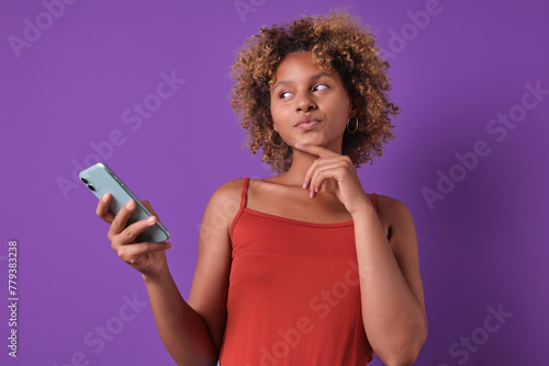 Young smiling casual African American woman teen with mobile phone in hand touches chin and looks to side thinking about message or post on social network stands on plain purple background.