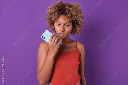 Young beauty embarrassed African American woman teen with phone in hand looks at screen thinking about advertising offer dressed in casual style stands on isolated purple background.