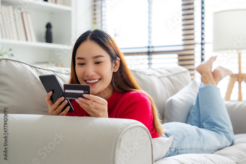 Young asian woman lying on sofa in living room, makes online banking payments through the internet from bank card on cell phone. Shopping online on mobile with credit card © Monster Ztudio