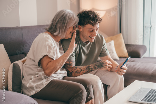 Video call concept. Happy senior retired woman sitting on sofa with young nephew waving hands video calling by mobile phone using technology for online webcam connection with distant family or friends photo
