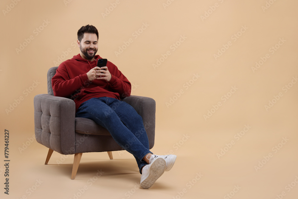 Happy young man using smartphone on armchair against beige background, space for text Happy young man using smartphone on armchair against beige background, space for text