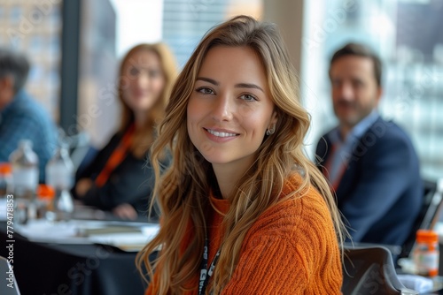 A woman with layered brown hair in a bright orange sweater is smiling at a formal event, sitting at a table with other people engaging in a fun conversation