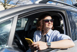 Smiling Caucasian woman in sunglasses sitting in a car in the parking lot driver's seat looking at the smartphone