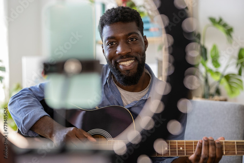 Happy influencer playing guitar and filming through smart phone at home photo