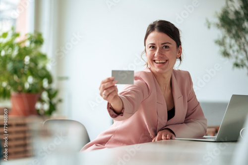 Happy businesswoman sitting with laptop at table showing business card working at home photo