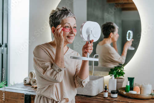 Smiling woman applying eye mask looking in mirror at home