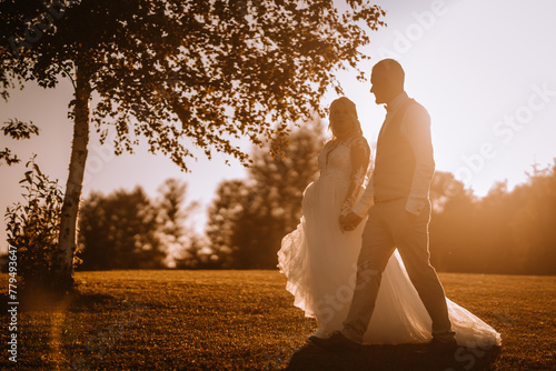 Valmiera, Latvia - Augist 13, 2023 - Silhouettes of a bride and groom holding hands against a sunset, with trees and a soft-focus background. photo