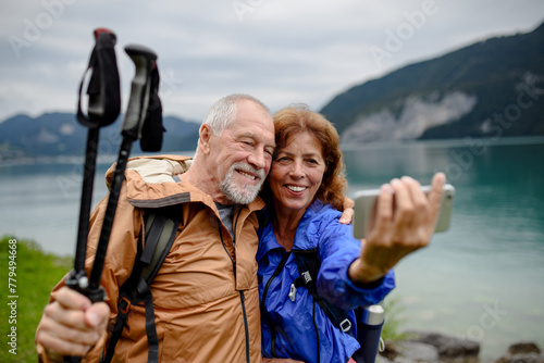 Portrait of beautiful active elderly couple taking selfie during hike in autumn mountains. Senior tourists embracing each other in front of lake © Halfpoint
