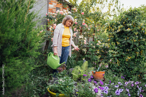 An elderly smiling woman waters flowers in hanging pots from a watering can