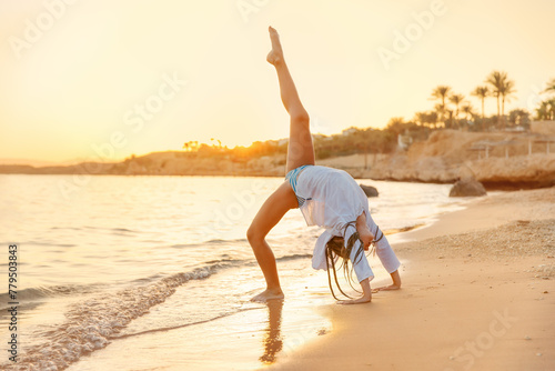 A girl is doing acrobatic exercises on the beach at sunset. She is standing on his hands in the sand, with his legs up in a split.