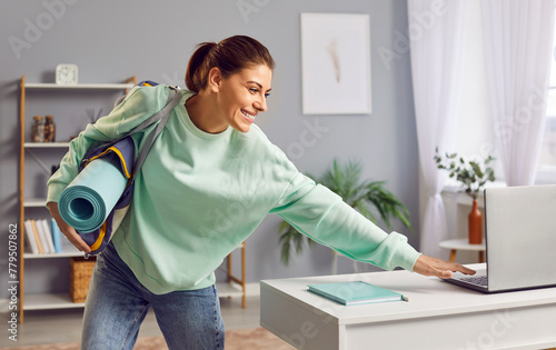 Positive woman with a sports bag, ready for training, turns off laptop on table, hurrying to leaving from home. She smiles, eager for gym session or outdoor workout, with fitness gear in bag.