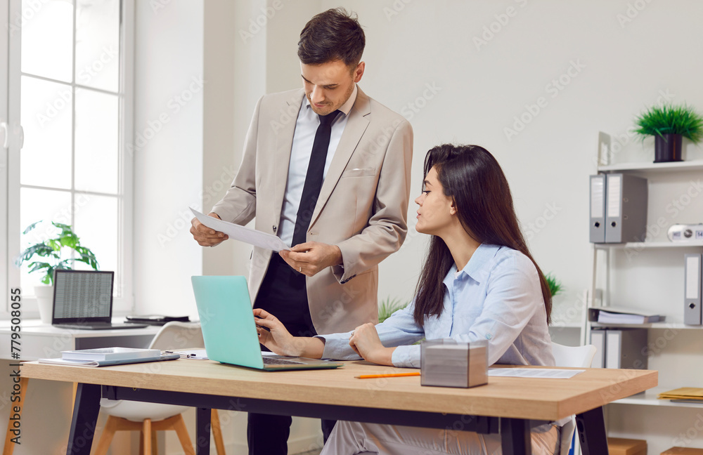 Two concentrated serious business people and company employees work in office sitting at the desk with laptop and looking through financial documents on workplace. Accountant job concept. Two concentrated serious business people and company employees work in office sitting at the desk with laptop and looking through financial documents on workplace. Accountant job concept.