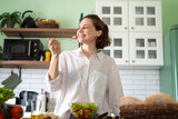 Young cheerful woman eating fresh vegetable salad while standing in the kitchen at home, healthy eating concept
