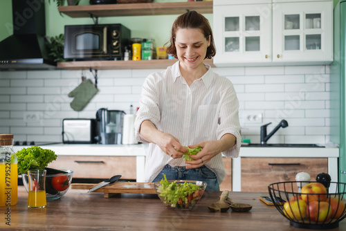 Young Caucasian woman cooking salad from green fresh vegetables while standing in the kitchen at home