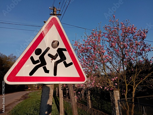 A triangular road sign depicting children running across the road. Children on the road sign on the background of a blossoming tree. photo