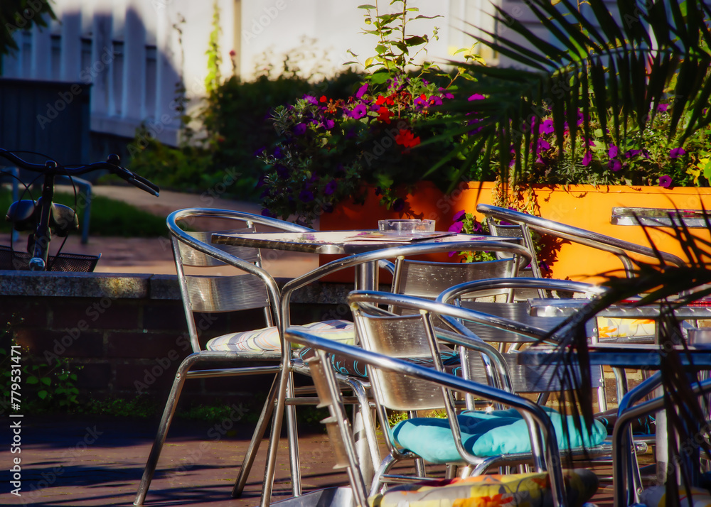 Im Vordergrund erstrecken sich die Stühle und Tische eines gemütlichen Cafés, liebevoll arrangiert, um die ersten Gäste in der sanften Morgenbrise zu empfangen. Die wärme der Sonnenstrahlen Im Vordergrund erstrecken sich die Stühle und Tische eines gemütlichen Cafés, liebevoll arrangiert, um die ersten Gäste in der sanften Morgenbrise zu empfangen. Die wärme der Sonnenstrahlen