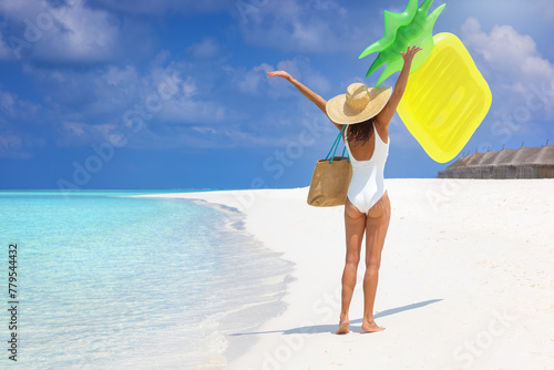 A happy woman in a white bathing suit walks down a tropical beach in the Maldives throwing a yellow floatie in the air