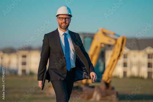 Successful construction business owner. Construction worker in suit and helmet near excavator. Confident construction owner in front of construction site. Civil engineer worker.