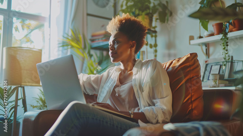 Middle-aged black woman sitting on the couch with a laptop on her knees with a thoughtful expression. Entrepreneurship, digital life and home.