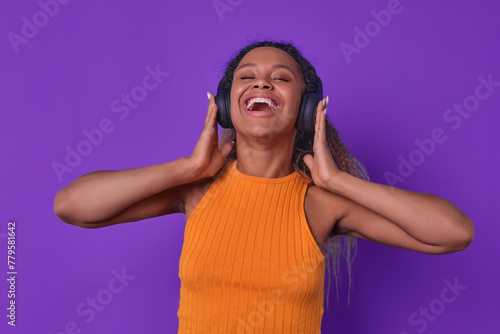 Young excited African American woman laughs pressing wireless headphones to ears listening to humorous audio podcast from comedians telling jokes or funny stories stands on purple background.