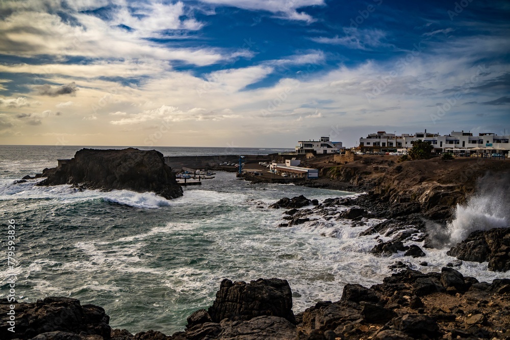 Beautiful shot of rugged rocks,the wavy ocean of El Cotillo in Spain and buildings in the background Beautiful shot of rugged rocks,the wavy ocean of El Cotillo in Spain and buildings in the background