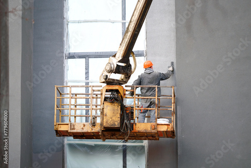 Worker on crane plastering building facade wall, construction worker in hardhat, work at height. Revamping building exteriors, stucco application by skilled worker and crane operation