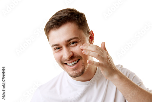 Young man in T-shirt applying cosmetic organic facial-care product smiling looking at camera against white background. Concept of beauty, facial-care, anti-aging, spa procedures, cosmetic products. photo